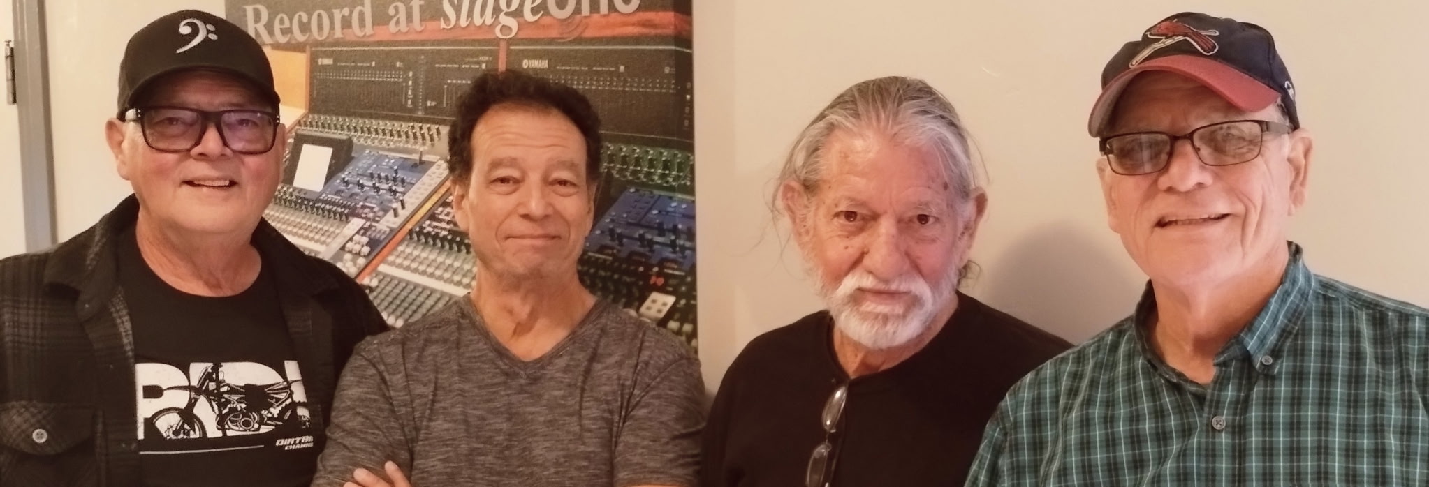The four members of Fast & Loose standing side-by-side in front of a poster for a recording studio.